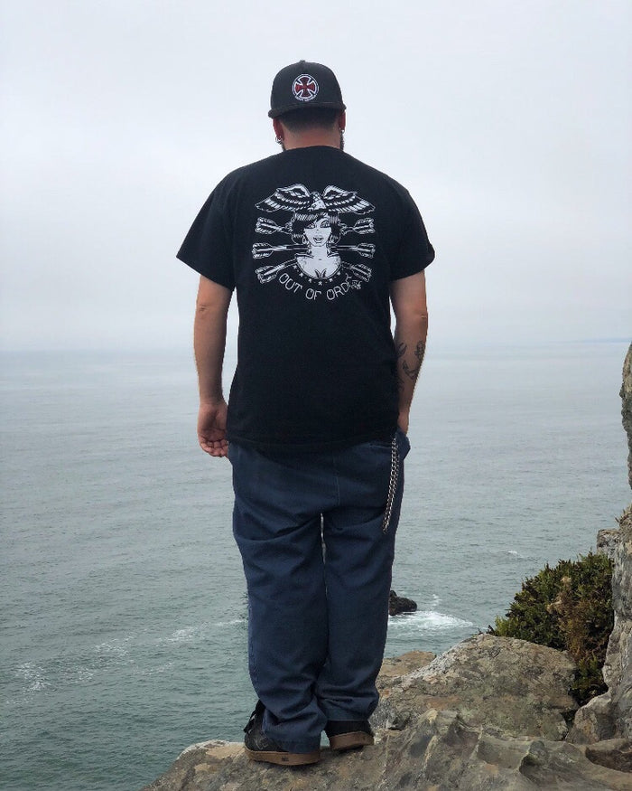 Male wearing tattoo style lady head shirt peering off cliff at ocean.